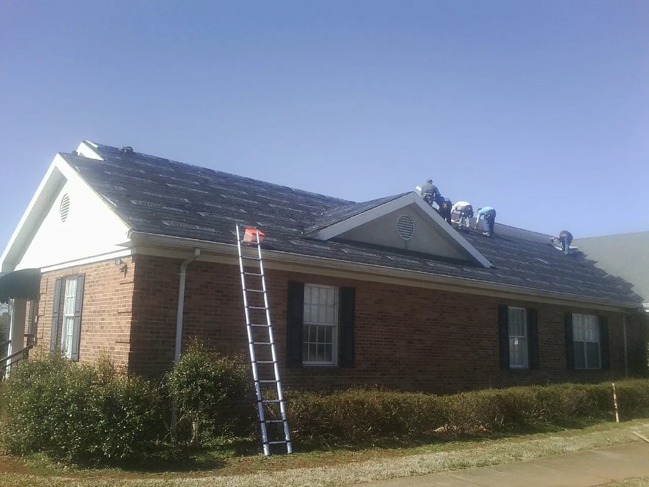 Crew tearing shingles off a brick ranch roof with a ladder set — Emergency Roof Repair work by our crew in George